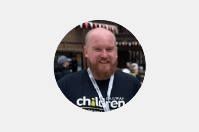 A man with a shaved head and brown beard wearing a black Railway Children T-shirt and a white Railway Children lanyard, smiling at a train station in front of some bunting.