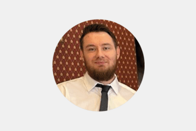 A man with dark hair and a dark beard wearing a white shirt and a black tie in front of a brown wall, smiling at the camera.