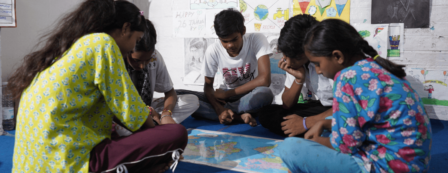 Children in India sat on the floor of a child activity centre classroom working and looking down at educational materials.