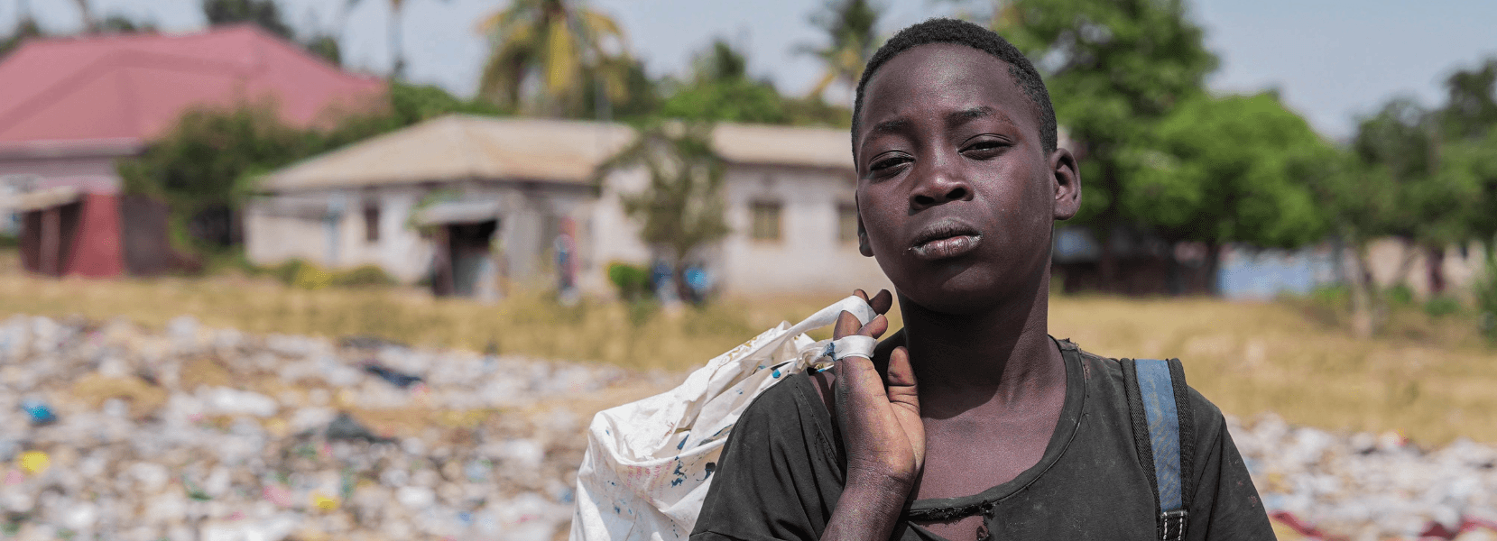 A young boy wearing a dark t-shirt carries a sack over his shoulder while standing in front of a rubbish-strewn area, with houses and palm trees in the background.