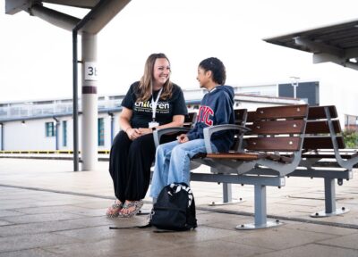 A Railway Children youth practitioner in a Railway Children black T-shirt and black trousers, wearing a lanyard, sat on a bench at a train station with a young teenage girl dressed in blue with a bag next to her.