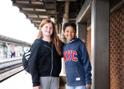 Two children at a train station in the UK with their arms round each other and smiling at the camera, one has red hair and is wearing black and grey and the other has dark hair and is dressed in blue.