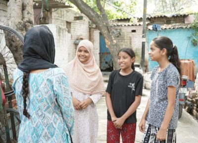 Three girls stand outdoors in a courtyard, smiling and talking with a Railway Children India project worker who has her back to the camera. The adult wears a patterned dress and a black headscarf, while the girls wear a mix of casual clothes and a hijab. Small houses, trees, and a motorbike are visible in the background.