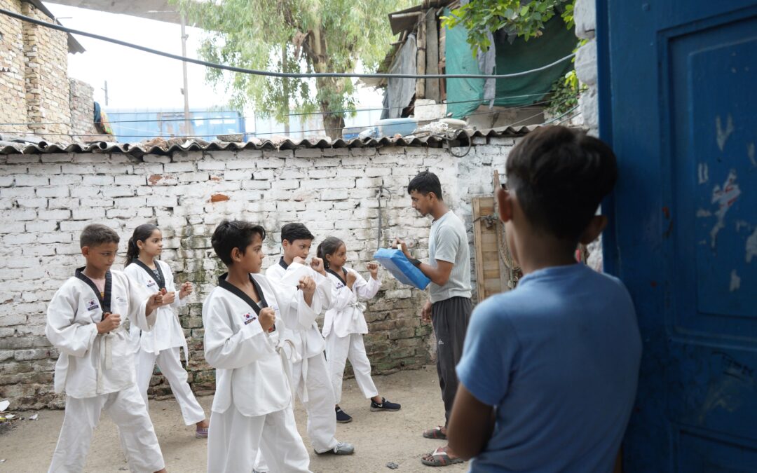 A boy wearing blue stood next to a blue door watching a group of children dressed in white learn karate with a teacher.