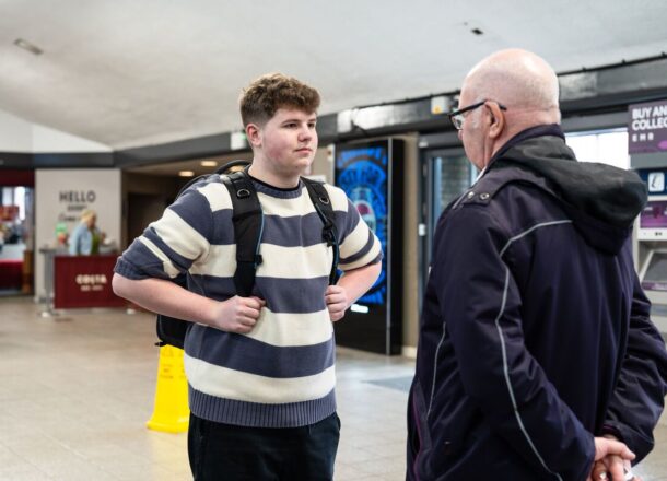 An older teenage boy with a blue and cream striped jumper, dark trousers and red hair carrying a rucksack at a train station in the UK talking to a member of the rail community.