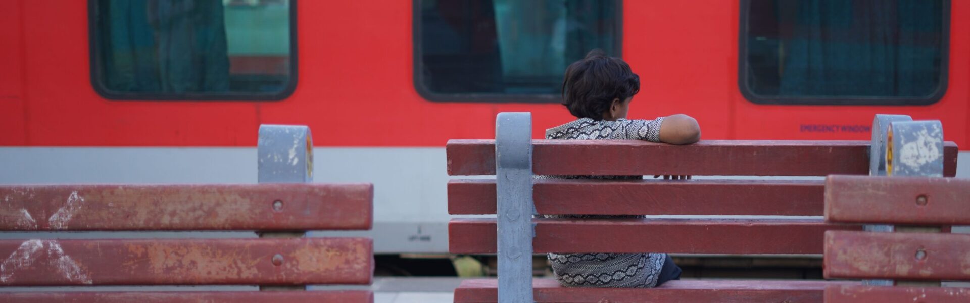 A young teenage boy sat on a bench at a station in India in front of a red train that is passing the platform.