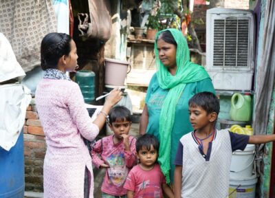 A mother wearing green and blue is stood behind her three children on the streets of India talking to a Railway Children project worker.