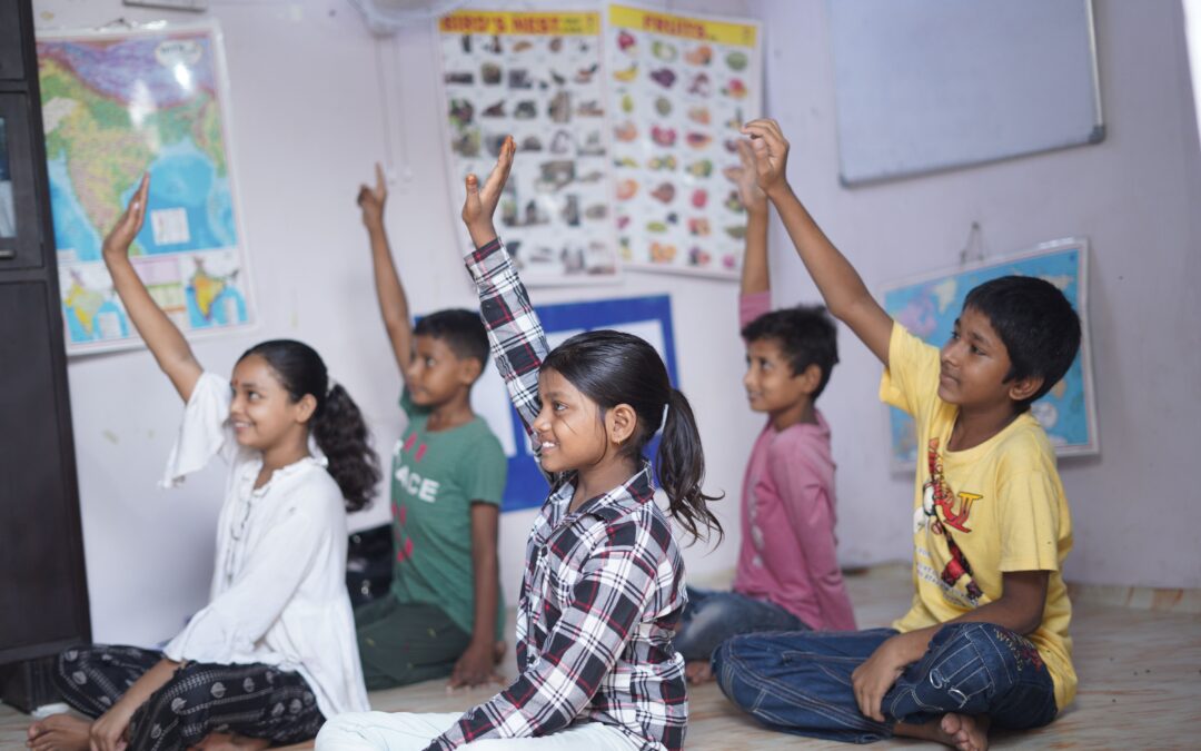 A group of children with their arms in the air sat on the floor addressing a teacher at a Child Activity Centre in India.