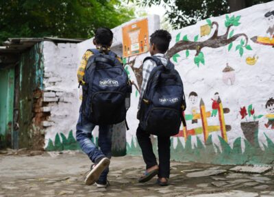 Two young boys carrying Railway Children branded backpacks, walking through their community in India.