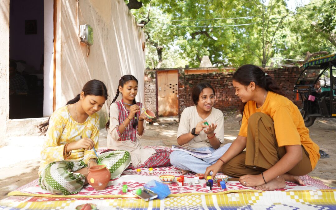 A group of girls sat outside on a blanket talking and painting some pottery various different colours, they all are looking happy and dressed in a variety of colours. There's a tree in the background and a brick wall.