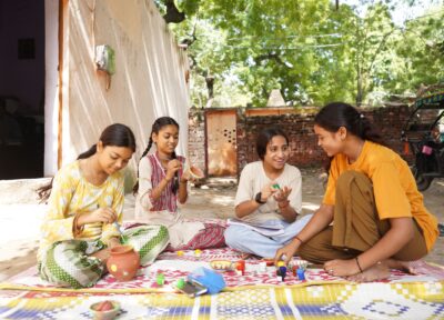 A group of girls sat outside on a blanket talking and painting some pottery various different colours, they all are looking happy and dressed in a variety of colours. There's a tree in the background and a brick wall.
