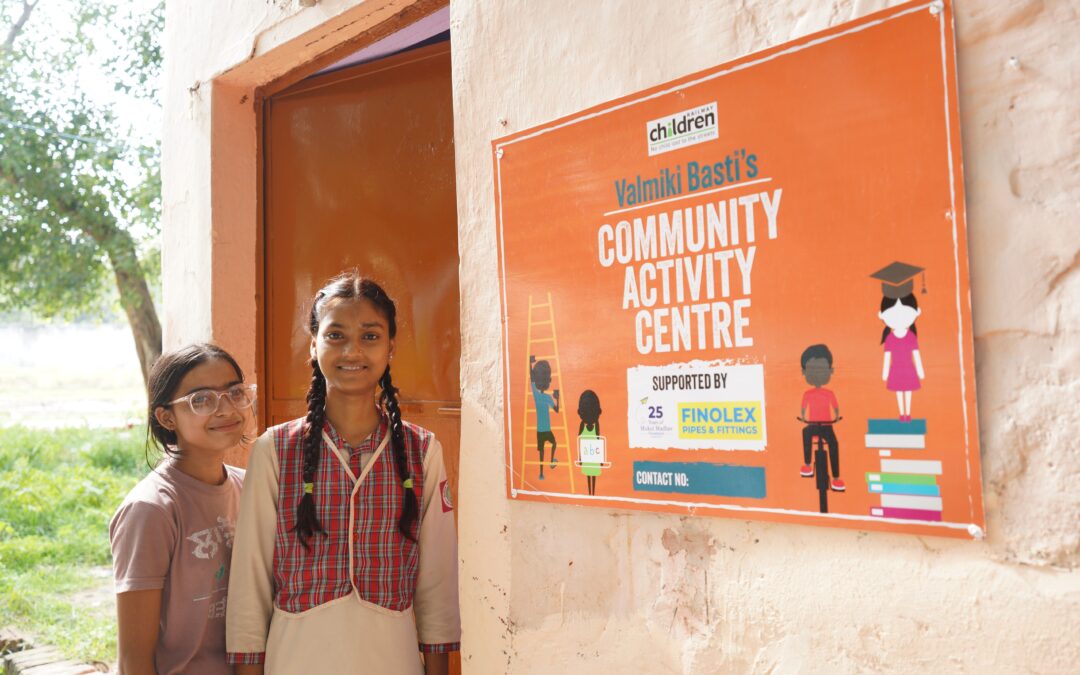 Two young girls stood next to a Child Activity Centre in India with an orange sign which reads Community Activity Centre.