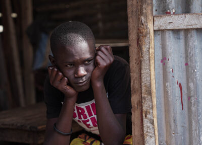 An older teenage boy looking out from behind a blue painted doorway wearing a black and pink T-shirt and holding his hands up to his face.
