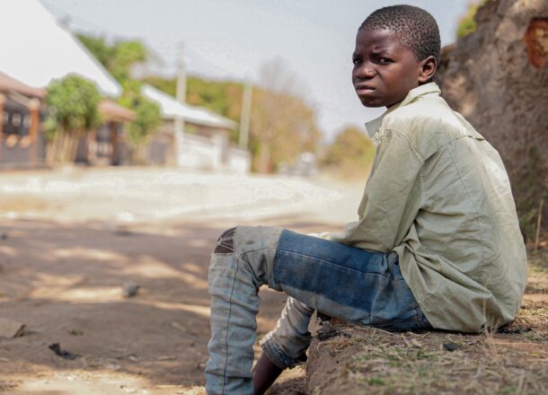 A young teenage boy in Tanzania with short hair sat on a step wearing a light coloured shirt and ripped jeans, looking out to the distance with a sad look on his face.