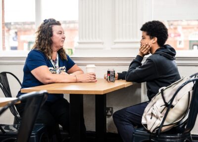 A Railway Children youth worker sits at a café table with a young person, having a supportive conversation. Both have drinks in front of them, and the atmosphere is calm and relaxed.