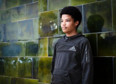 A younger teenage boy with dark short hair looking out to the distance in front of a green tiled wall and wearing a black tracksuit top.
