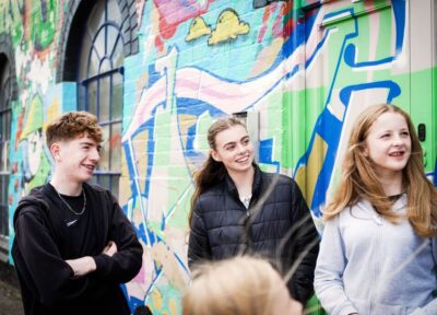 A teenage boy with dark blonde hair wearing a black coat and two blonde teenage girls wearing black and grey hooded jackets stood in front of a colourful graffiti wall looking to the right and smiling.