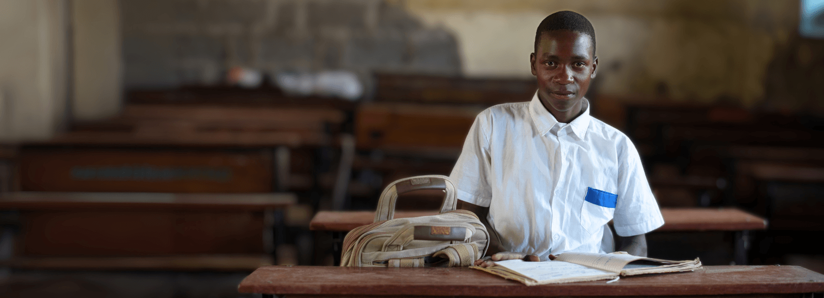 An older teenage boy in a white shirt sat in a dark classroom at a desk with a notepad and backpack.
