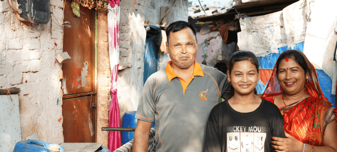 A teenage girl wearing a black T-shirt stood in the middle of her mother (wearing orange) and her father who is wearing a grey and orange polo neck top. They are all smiling and stood in the street next to a home.