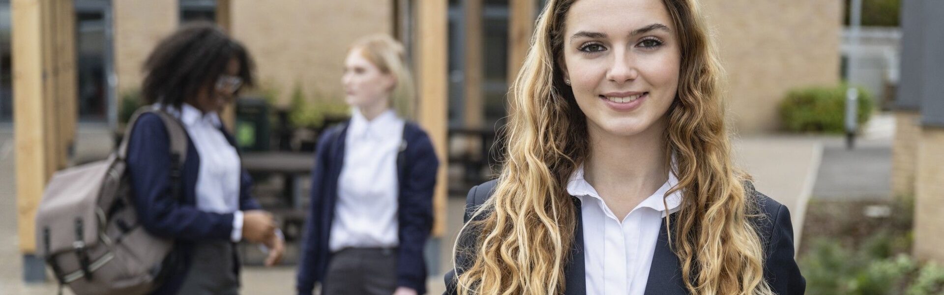 A teenage girl in the UK holding a backpack and wearing school uniform with some friends in the background.
