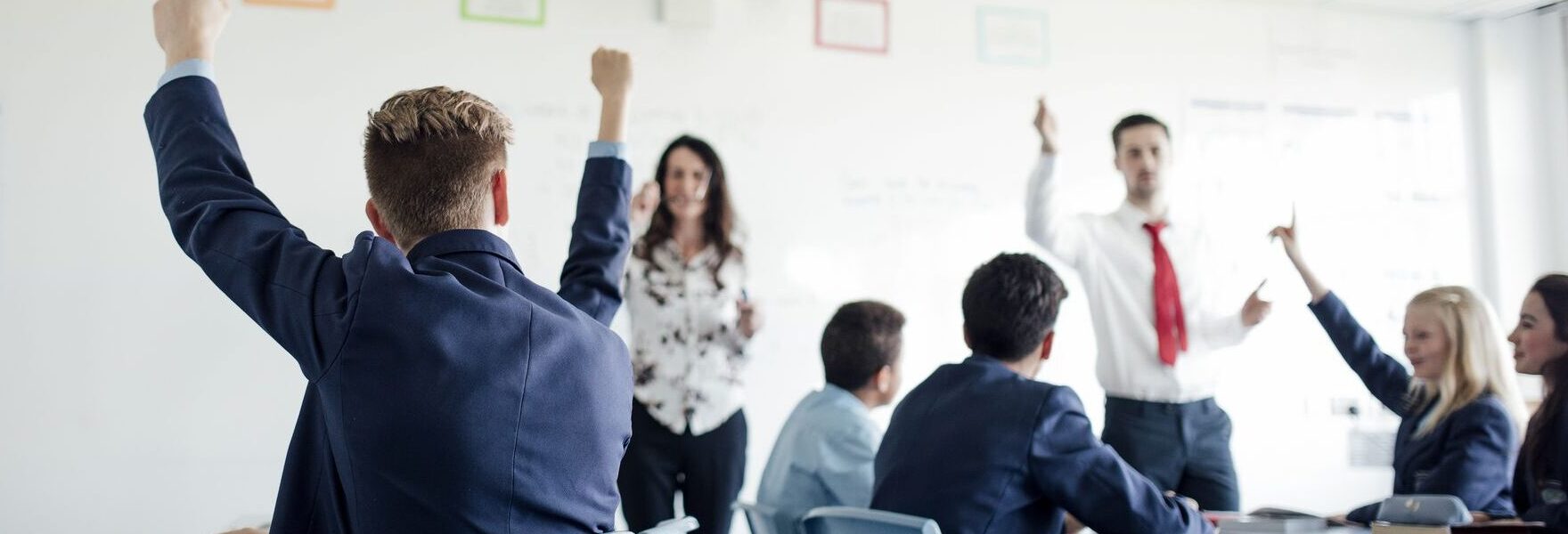 A classroom with teenagers sat at desks with their arms up in the air and two teachers stood at the front next to whiteboards with drawings on them.