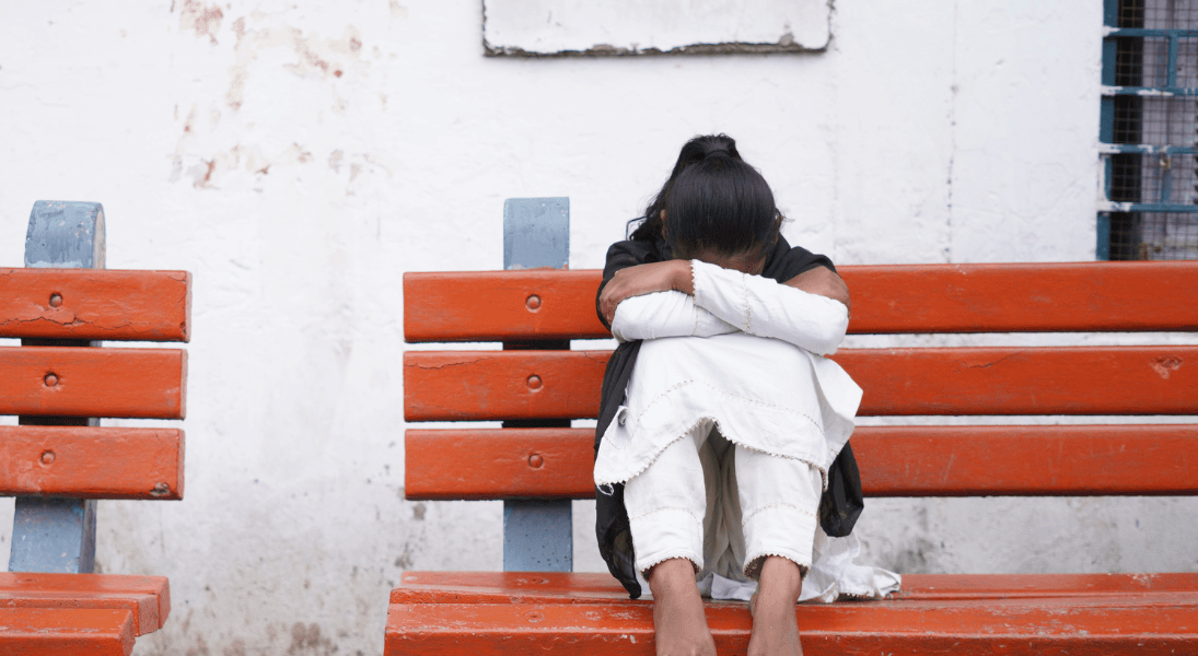 A girl sat on a bench with her legs up, leaning her arms into her legs and her face down.