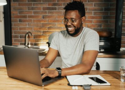 A man sat in a kitchen in front of a brick patterned wall wearing glasses and a grey T-shirt working on a laptop on a wooden kitchen counter and smiling.