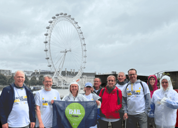 A group of participants stand together outdoors during the Railway Children Rail Trail event in London. They are smiling at the camera, some wearing event t-shirts and rain ponchos, while holding a Rail Trail banner. Behind them, the London Eye is visible against a cloudy sky.