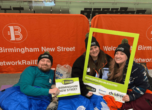 Three people sit close together in sleeping bags at a Railway Children Sleepout event. They are smiling at the camera and holding a large yellow photo frame with the Railway Children Sleepout branding and website details. The event is taking place inside a train station, with metal barriers and signage visible in the background.