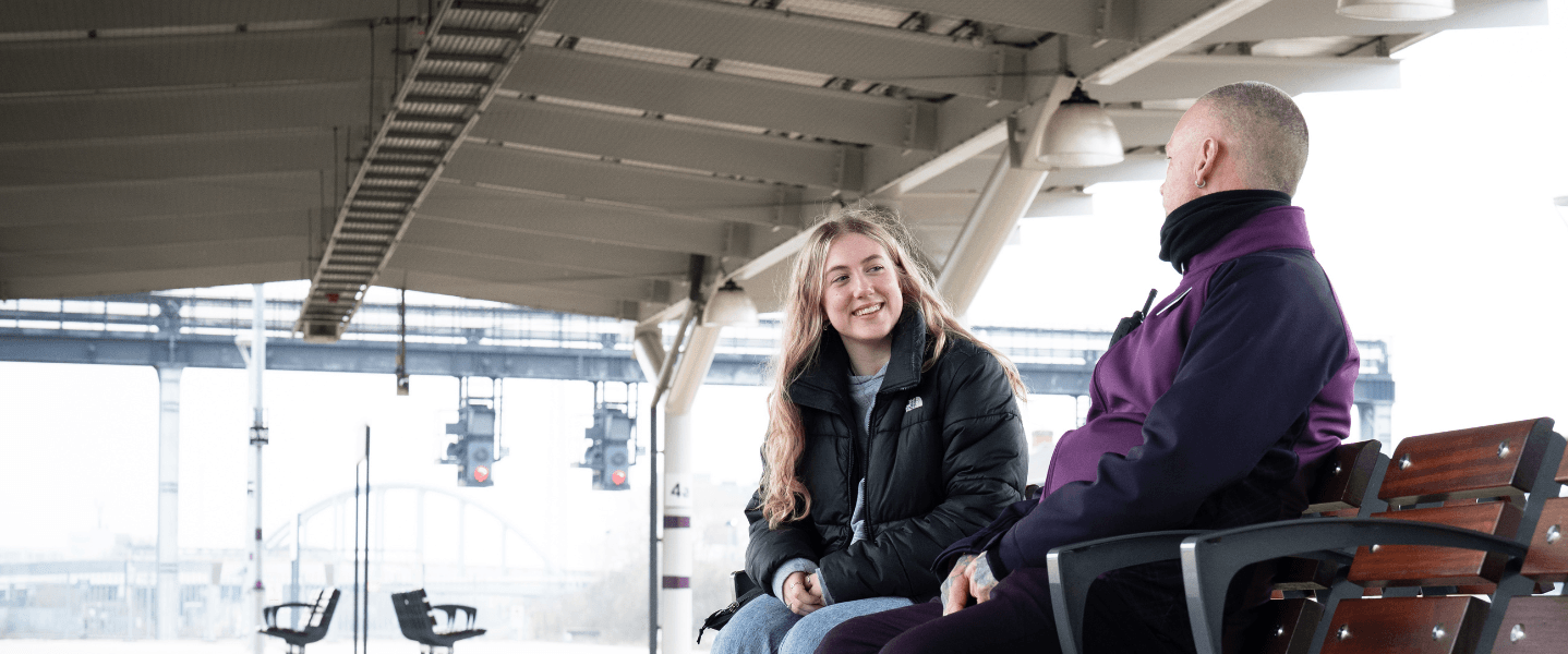 A blonde teenage girl wearing a black coat and jeans sat on a bench on the platform of a UK rail station talking to a member of rail staff wearing a purple jacket.
