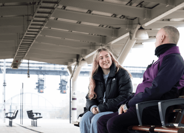 A blonde teenage girl wearing a black coat and jeans sat on a bench on the platform of a UK rail station talking to a member of rail staff wearing a purple jacket.