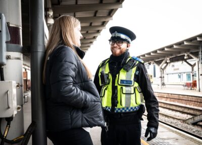A teenage girl with red hair with her hands in the pockets of her dark jacket, leaning against a wall and talking to a British Transport Police officer who is smiling, next to rail tracks on a platform.