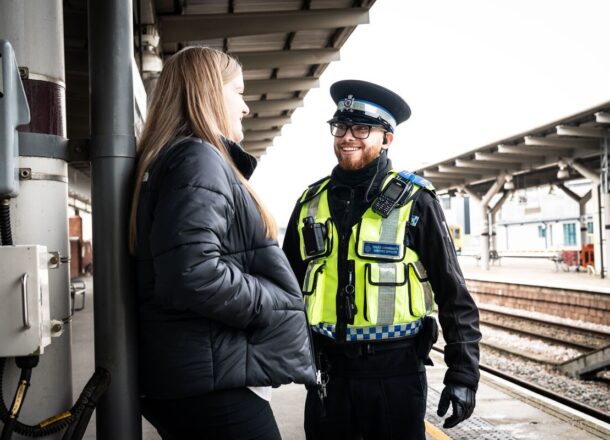A teenage girl with red hair with her hands in the pockets of her dark jacket, leaning against a wall and talking to a British Transport Police officer who is smiling, next to rail tracks on a platform.