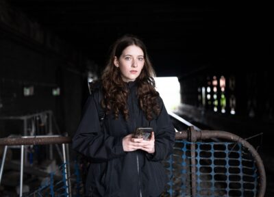A teenage girl with long brown hair and blue eyes stands under a dark railway bridge, wearing a black jacket and holding a mobile phone. The background is blurred, showing the tunnel and light at the far end, creating a contrast that highlights her serious expression.