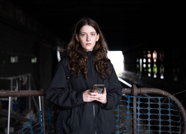 Teenage girl with long brown hair wearing a black jacket, standing under a dark railway bridge holding a phone, symbolising vulnerability and awareness of youth issues.
