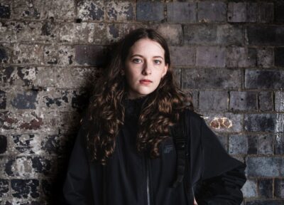 A teenage girl with brown hair wearing black stood in front of a grey brick wall looking serious and at the camera.