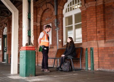 A member of staff from the rail industry wearing an orange hi-vis jacket stood next and talking to a teenage girl who is wearing dark clothing and sat on a bench at a railway station in the UK.