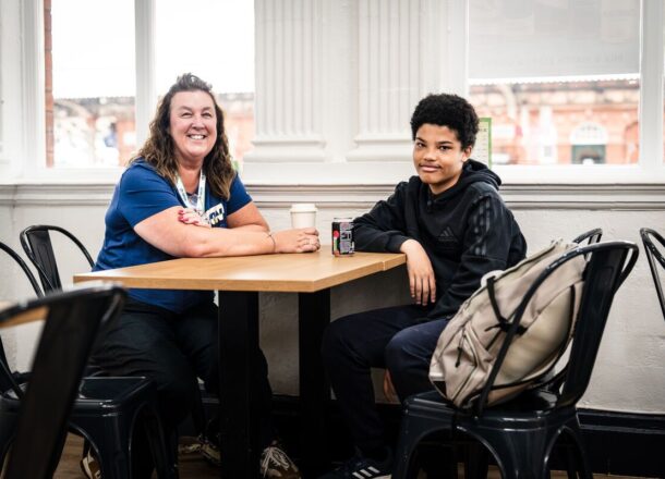 A Railway Children youth worker sits at a café table with a young person, they are both leaning on the table and smiling, looking at the camera.