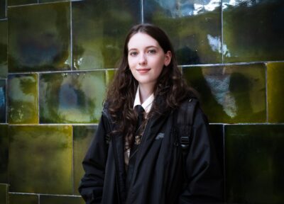 A teenage girl wearing school uniform and a black coat with dark brown hair stood in front of a green tiled wall and smiling at the camera.