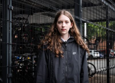 A teenage girl with brown hair and pale skin wearing a dark coloured jacket, smiling slightly in front of some railings.