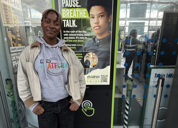 A Railway Children youth ambassador wearing a grey hoodie which reads Youth Platform standing next to one of Railway Children's screens at a train station in the UK smiling and standing proudly.