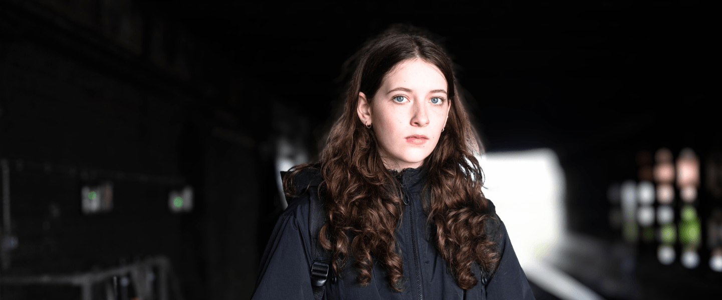 A teenage girl with long brown hair and blue eyes stands under a dark railway bridge, wearing a black jacket and holding a mobile phone. The background is blurred, showing the tunnel and light at the far end, creating a contrast that highlights her serious expression.