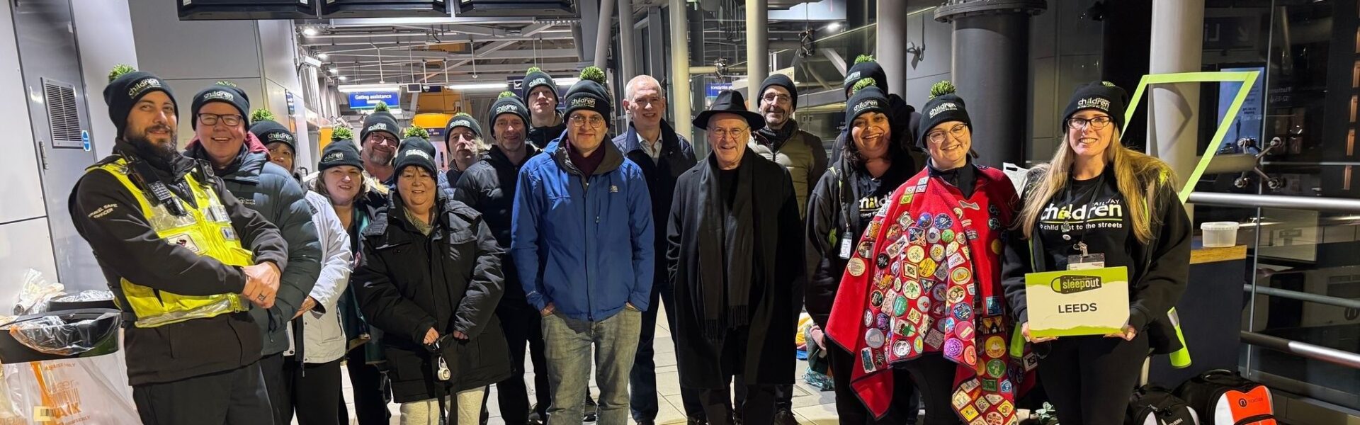 A group of Railway Children charity supporters and participants gather at Leeds Station for the Railway Children Sleepout event. Everyone is warmly dressed in coats and Railway Children beanies, smiling as they prepare to spend the night raising awareness and funds for vulnerable children. One person holds a sign reading "LEEDS," and another wears a badge-covered red blanket. The event takes place on the station platform, supported by volunteers and rail staff.