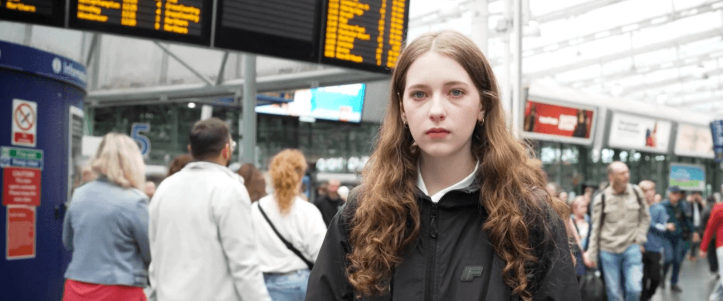 Young teenage girl with brown hair and pale skin wearing a dark jacket stood in front of crowds of people at a railway station in the UK.