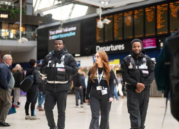 A Railway Children project worker wearing a black Railway Children T-shirt and lanyard walking through Waterloo Station with two security officers wearing black walking either side of her.