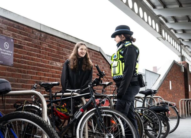 A teenage girl stood next to some bikes parked at a station talking to a female British Transport Police officer.