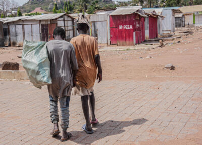 Two teenage boys walking through the streets of Tanzania wearing dirty clothes of various colours and carrying rubbish sacks.