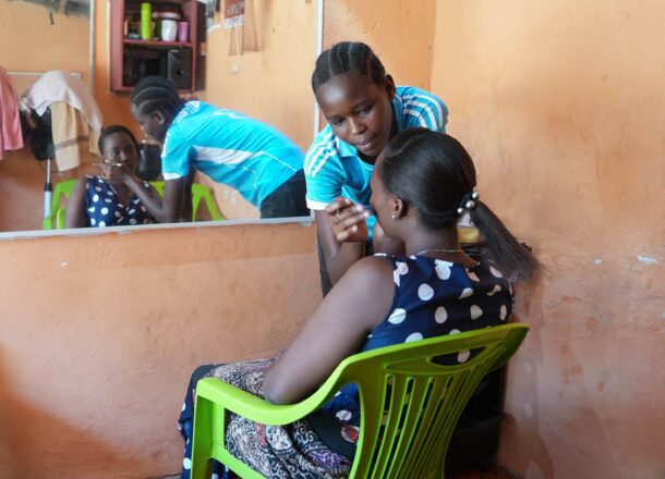 A young adult female wearing a blue T-shirt putting make up on a woman who is sitting in a green chair wearing a spotty navy and white dress in front of a mirror in a beauty salon in Tanzania.