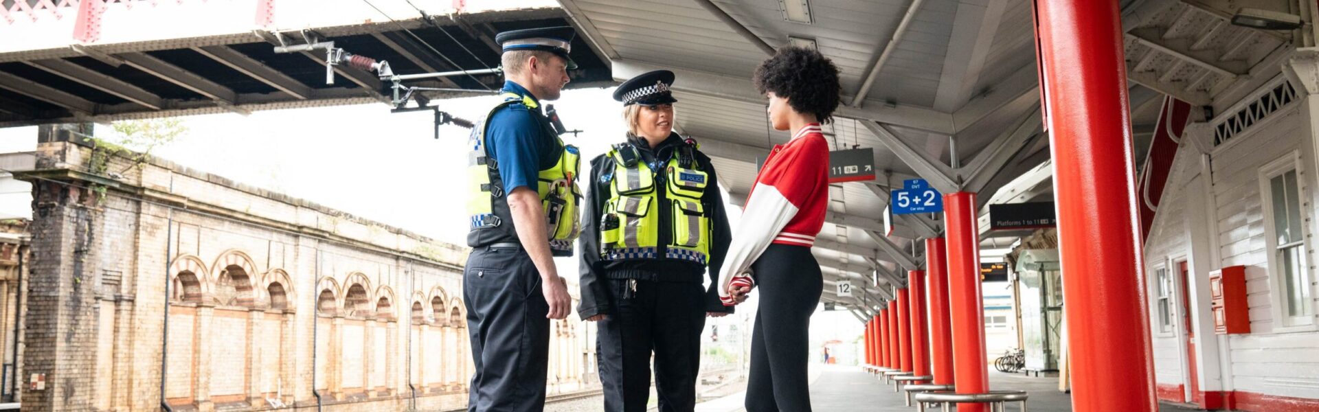 Two British Transport Police officers stood on the platform of a railway station in the UK next to the tracks talking to a young teenage girl wearing a red jacket and black leggings.