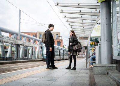 A young teenage girl talking to an older teenage boy stood on a platform at a train station in the UK.